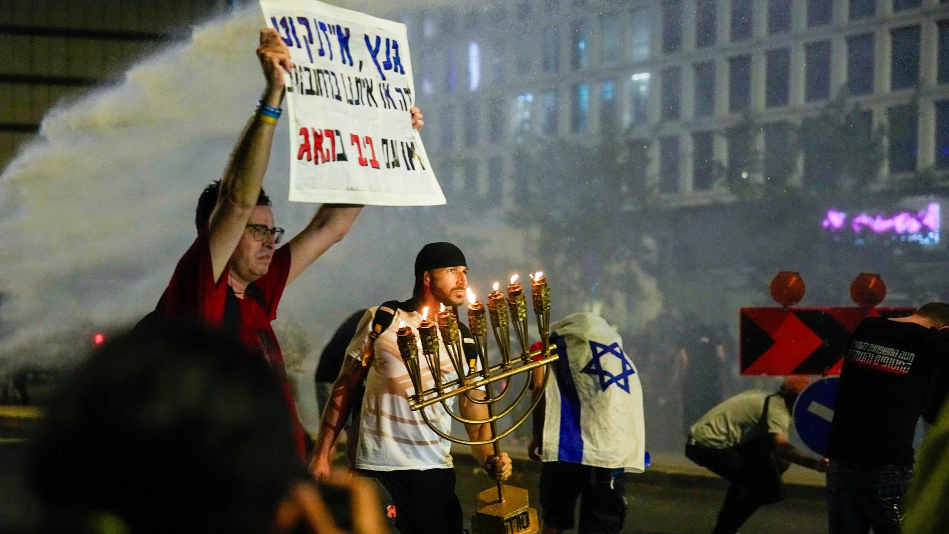 Proteste in Tel Aviv gegen die israelische Regierung und Premierminister Netanjahu. Ein Demonstrant hält ein Schild mit hebräischer Schrift in die Höhe, ein anderer hält einen Kerzenleuchter mit sieben Lichtern. 