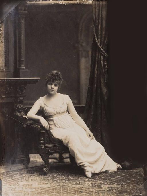 Fotografie der jungen russischen Künstlerin Maria Bashkirtseff, die auf einem Stuhl sitzt in einem weißen Kleid und hochgesteckten Haaren.