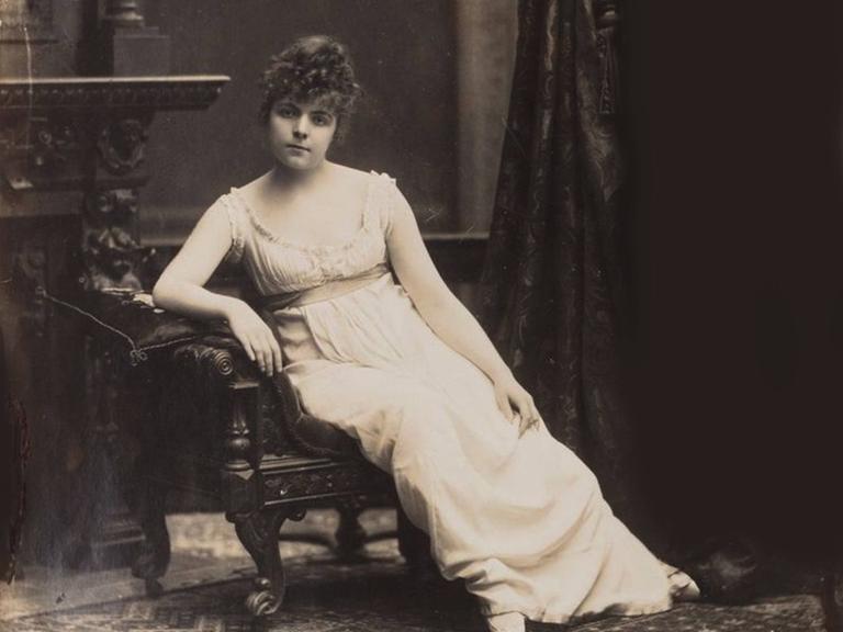 Fotografie der jungen russischen Künstlerin Maria Bashkirtseff, die auf einem Stuhl sitzt in einem weißen Kleid und hochgesteckten Haaren.