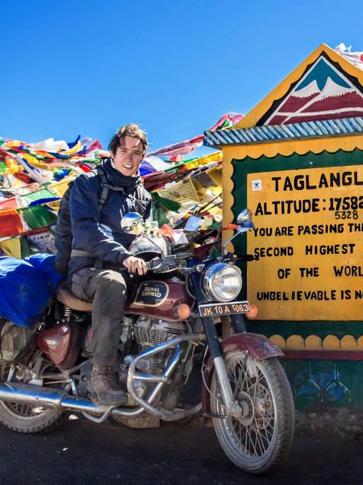 Erik Lorenz posiert auf einem Motorrad vor einem Schild, auf dem 'Taglangla' steht und an dem unzählige Schnüre mit bunten Gebetsfähnchen befestigt sind.