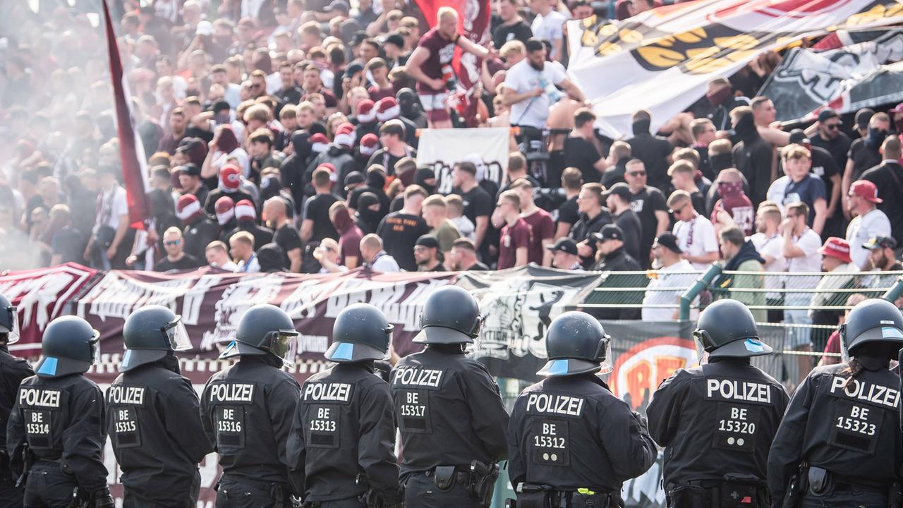 Gewaltbereite Fußballfans stehen auf einer Tribüne. Ein Großaufgebot an Polizisten sichert das Stadion. 