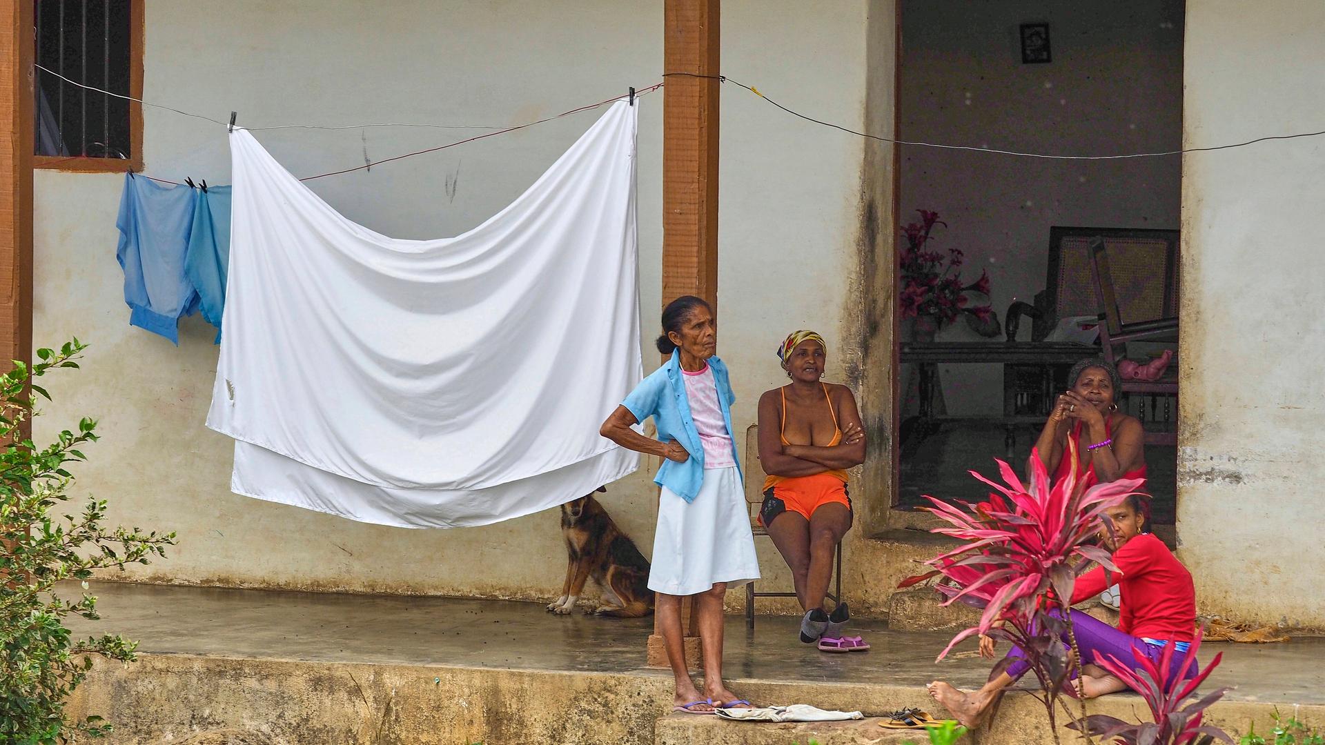 Vier kubanische Frauen halten sich vor einem Wohnhaus auf. Drei Frauen sitzen, eine steht. Ein Bettlaken hängt auf einer Wäscheleine. Im Hintergrund sitzt ein Hund. 