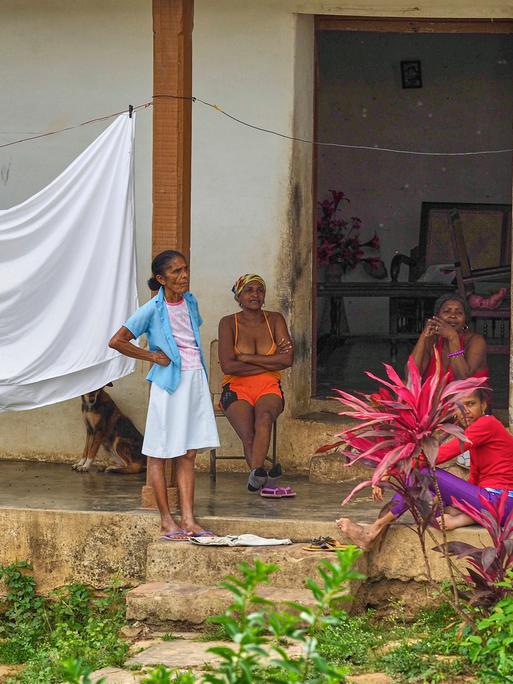 Vier kubanische Frauen halten sich vor einem Wohnhaus auf. Drei Frauen sitzen, eine steht. Ein Bettlaken hängt auf einer Wäscheleine. Im Hintergrund sitzt ein Hund. 