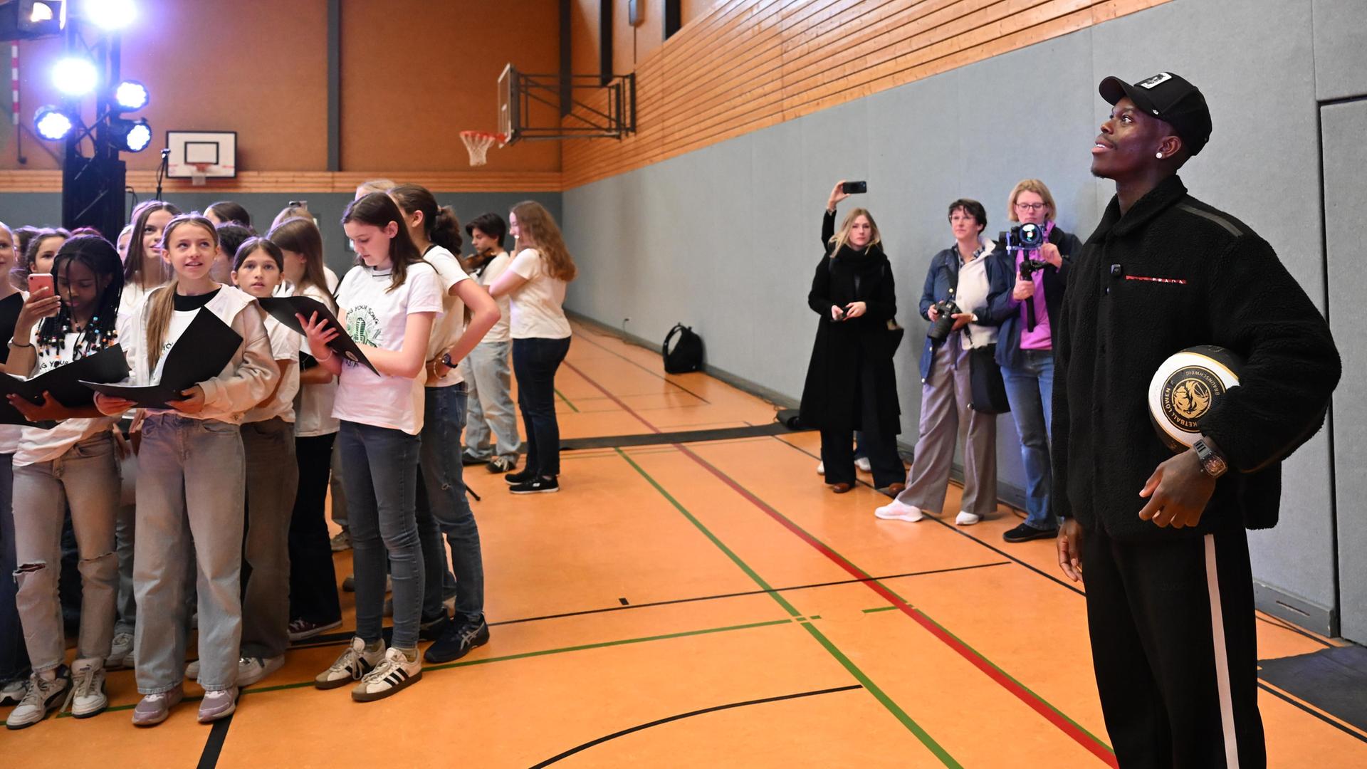 Ein schwarzer Mann mit Kappi und Basketball unterm Arm steht in einer Sporthalle.