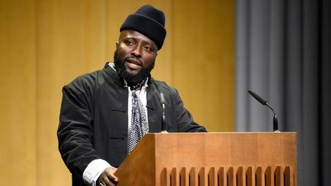 Bonaventure Soh Bejeng Ndikung spricht im Haus der Kulturen der Welt in Berlin an einem Pult vor einem Mikrofon.