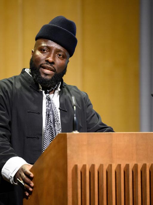 Bonaventure Soh Bejeng Ndikung spricht im Haus der Kulturen der Welt in Berlin an einem Pult vor einem Mikrofon.