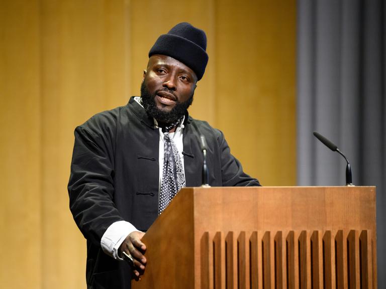Bonaventure Soh Bejeng Ndikung spricht im Haus der Kulturen der Welt in Berlin an einem Pult vor einem Mikrofon.