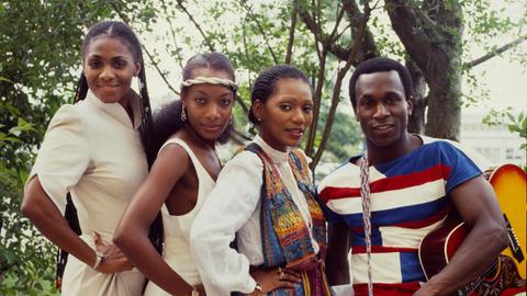 Die drei weiblichen und das männliche Mitglied der Band Boney M stehen dicht zusammen im Freien. Der Mann rechts hat eine Gitarre von der Schulter baumeln und hält sie mit einer Hand fest.