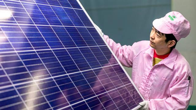 Ein chinesischer Mann hält ein großes Solarmodul in den Händen und schaut prüfend darauf. Er trägt einen rosafarbenen Arbeitsoverall und Mütze. Ein chinesischer Mann hält ein großes Solarmodul in den Händen und schaut prüfend darauf. Er trägt einen rosafarbenen Arbeitsoverall und Mütze.