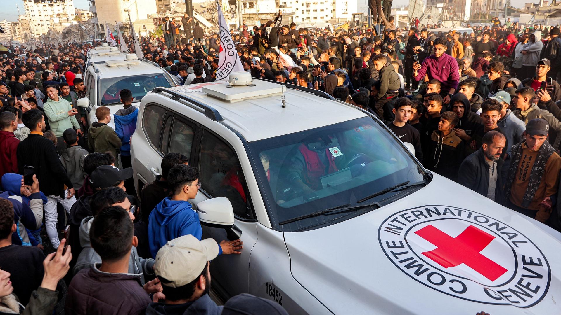 Mehrere Autos vom Roten Kreuz fahren durch eine Menschen-Menge.