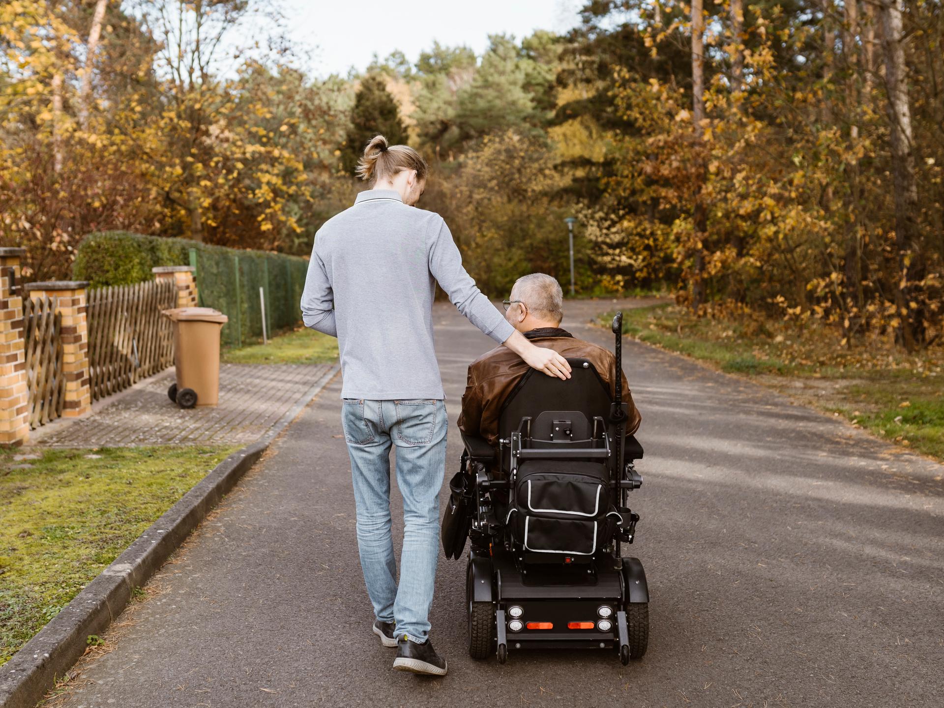 Eine junge Person läuft neben einer Person im Rollstuhl. Sie laufen bzw. fahren über eine ruhige Straße am Waldrand. (Symbolbild) Eine junge Person läuft neben einer Person im Rollstuhl. Sie laufen bzw. fahren über eine ruhige Straße am Waldrand. (Symbolbild)