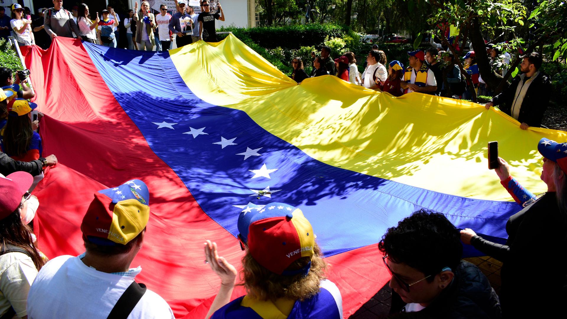 Dutzende Venezolanerinnen und Venezolaner halten gemeinsam eine große venezolanische Flagge. Dutzende Venezolanerinnen und Venezolaner halten gemeinsam eine große venezolanische Flagge.