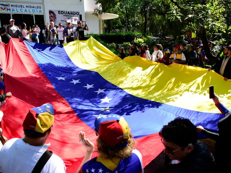Dutzende Venezolanerinnen und Venezolaner halten gemeinsam eine große venezolanische Flagge.