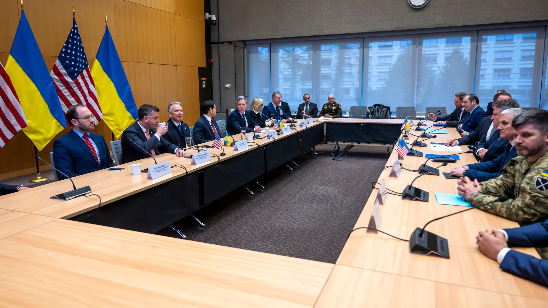 Mehrere Männer in Anzügen sitzen sich an zusammengestellten Tischen gegenüber. Im Hintergrund sind die Flaggen der USA und der Ukraine zu sehen.