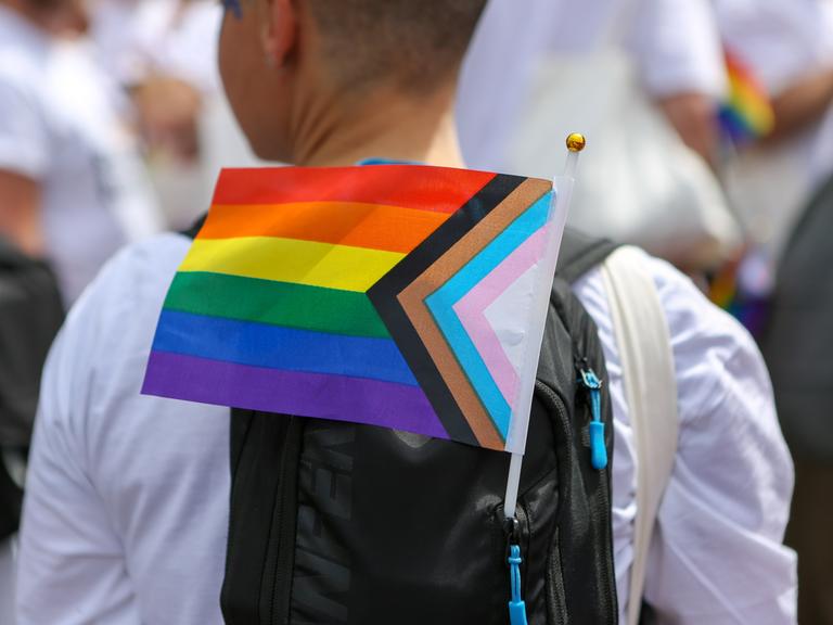 Eine Person nimmt mit einer Pride-Flagge am Rucksack an einer Demonstration teil.