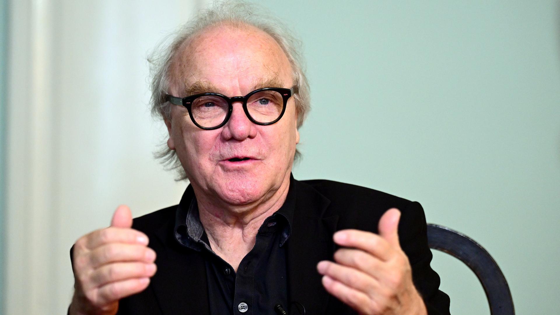 Autor Michael Köhlmeier sitzt an einem Tisch und blickt auf sein Gegenüber. Der Autor trägt eine schwarze Brille und Hemd. 