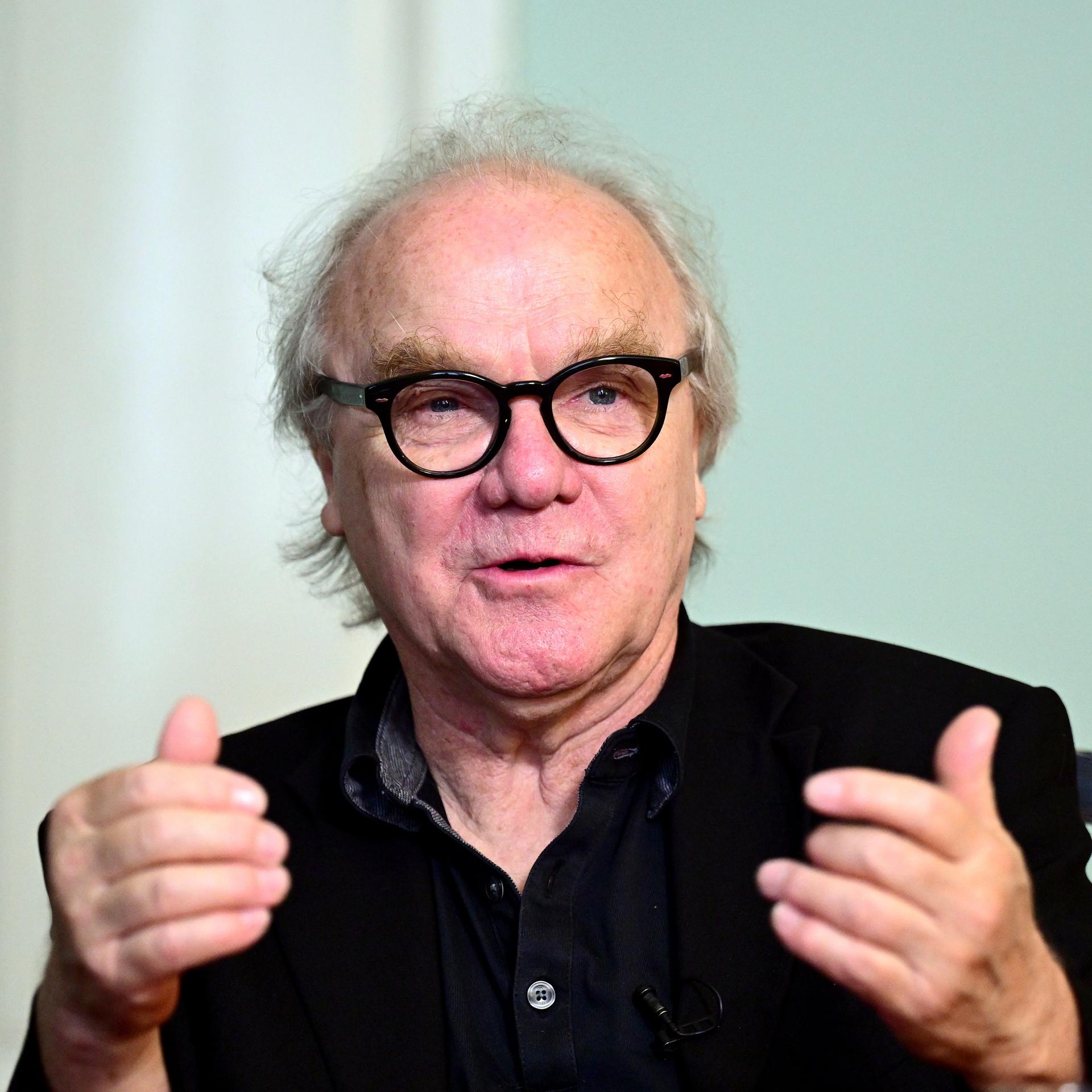 Autor Michael Köhlmeier sitzt an einem Tisch und blickt auf sein Gegenüber. Der Autor trägt eine schwarze Brille und Hemd. 