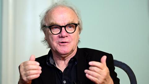 Autor Michael Köhlmeier sitzt an einem Tisch und blickt auf sein Gegenüber. Der Autor trägt eine schwarze Brille und Hemd. 