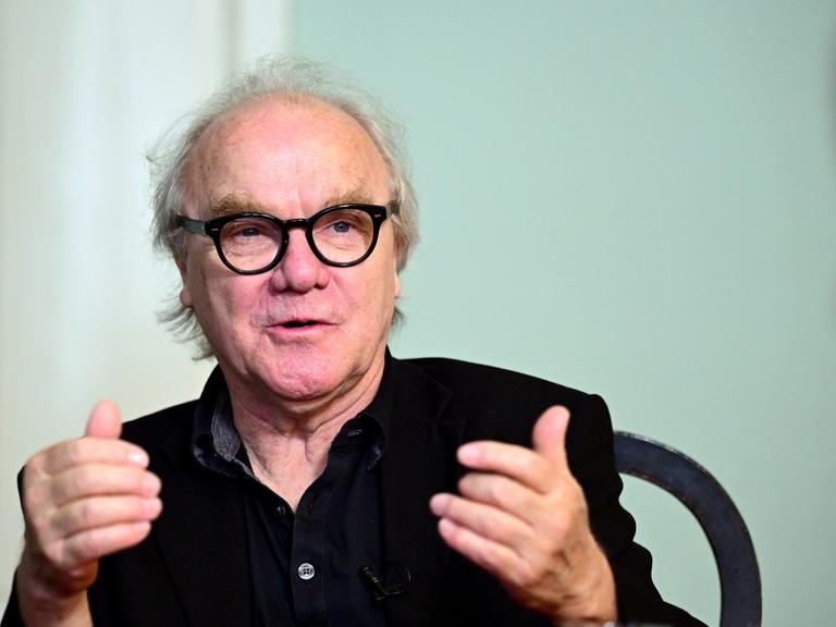 Autor Michael Köhlmeier sitzt an einem Tisch und blickt auf sein Gegenüber. Der Autor trägt eine schwarze Brille und Hemd. Autor Michael Köhlmeier sitzt an einem Tisch und blickt auf sein Gegenüber. Der Autor trägt eine schwarze Brille und Hemd.