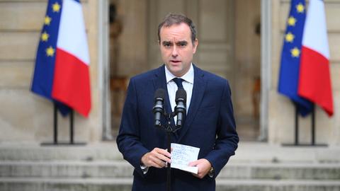 Zu sehen ist Sébastien Lecornu, wie er im Freien vor zwei Mikrofonen steht. Er trägt einen dunkelblauen Anzug und hält einen Zettel in den Händen. Im Hintergrund die Flaggen Frankreichs und der EU.
      