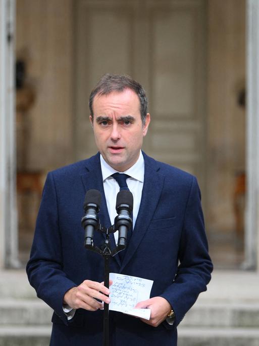 Zu sehen ist Sébastien Lecornu, wie er im Freien vor zwei Mikrofonen steht. Er trägt einen dunkelblauen Anzug und hält einen Zettel in den Händen. Im Hintergrund: die Flaggen Frankreichs und der EU. 
