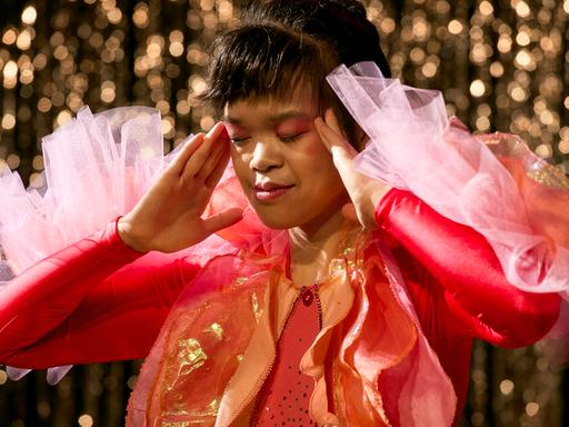 Eine Frau in rotem Kleid steht auf einer Theaterbühne und berührt mit ihren Händen die Schläfen.