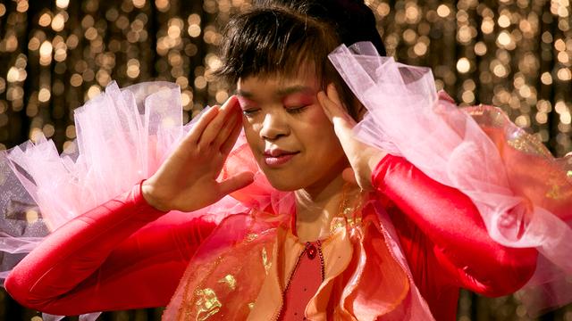 Eine Frau in rotem Kleid steht auf einer Theaterbühne und berührt mit ihren Händen die Schläfen.