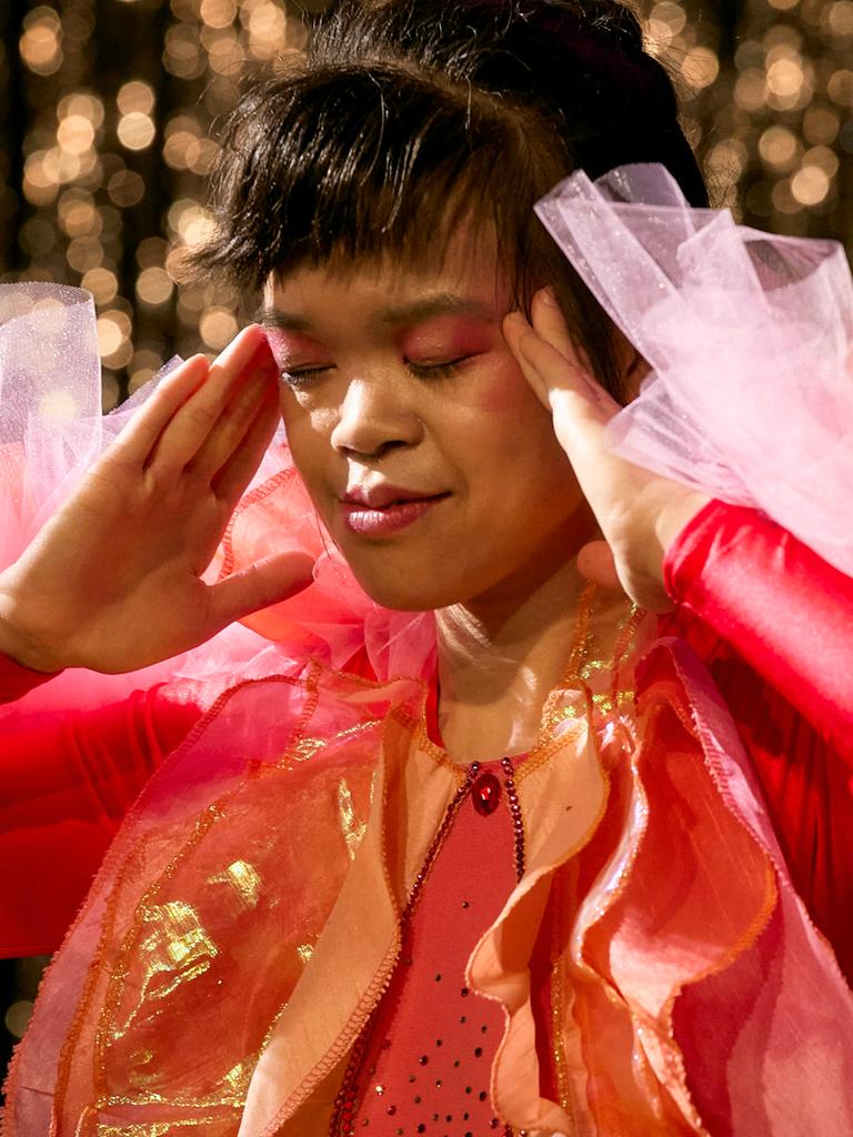 Eine Frau in rotem Kleid steht auf einer Theaterbühne und berührt mit ihren Händen die Schläfen.