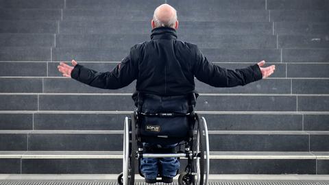 Ein Mann sitzt im Rollstuhl vor einer Treppe ohne Rampe und kommt nicht weiter.