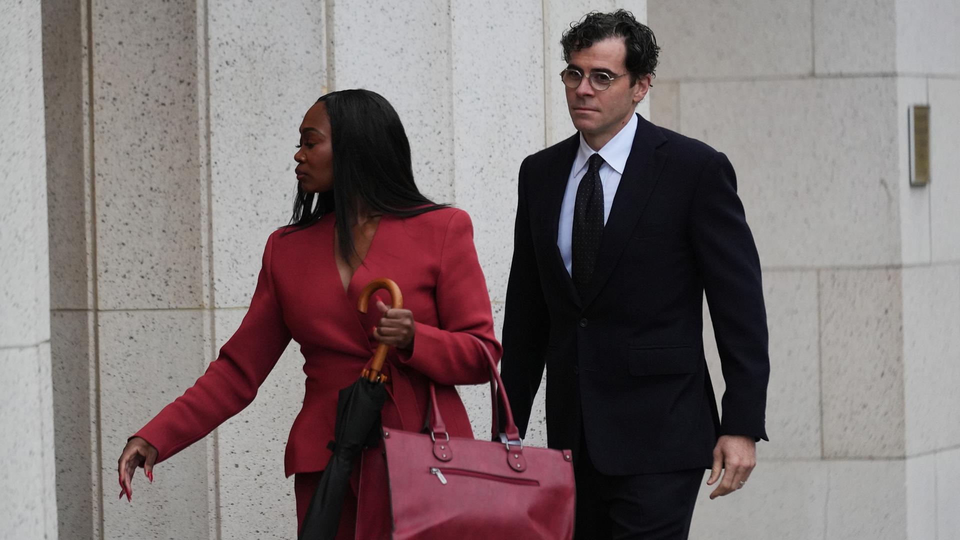 Mosseri mit Brille und dunklem Anzug und die in einem roten Kostüm gekleidete schwarze Anwältin gehen gerade zum Eingang der Gerichtsgebäudes. 