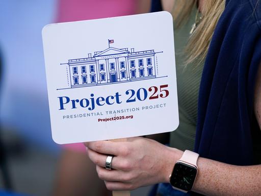 Eine Person hält ein Schild mit der Aufschrift "Project 2025" in der Hand. Darauf ist außerdem eine Zeichnung des Weißen Hauses zu sehen.