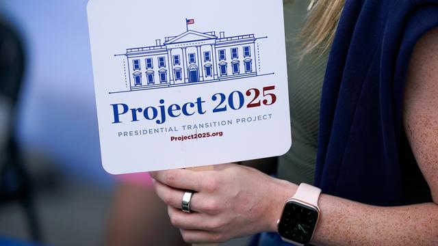 Eine Person hält ein Schild mit der Aufschrift "Project 2025" in der Hand. Darauf ist außerdem eine Zeichnung des Weißen Hauses zu sehen.