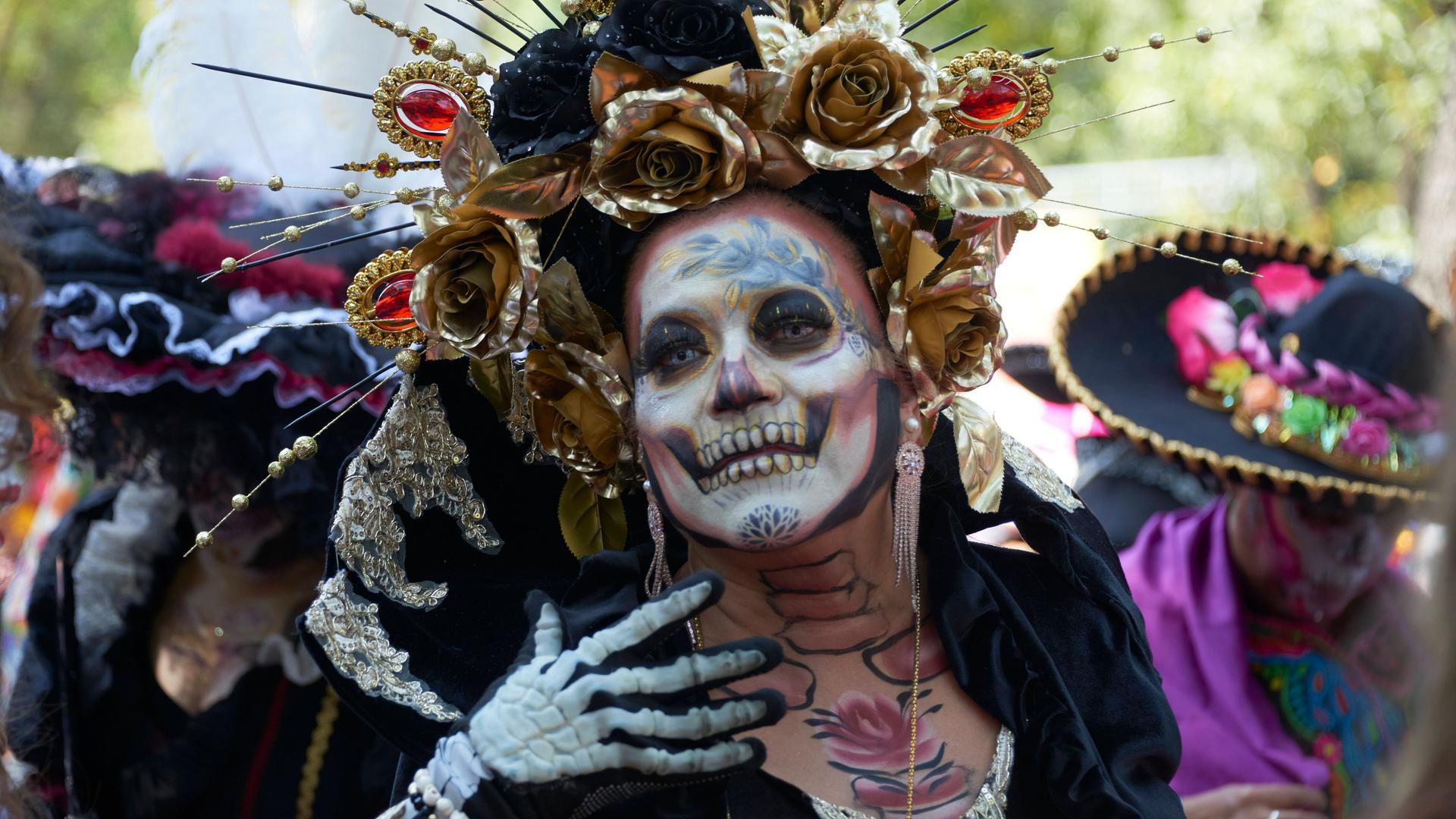 Eine Frau hat Gesicht und Hände als Skelett geschminkt, trägt einen großen, mit goldenen Rosen verzierten Hut und geht bei der Parade am 04.11.2023 in Mexico City mit.