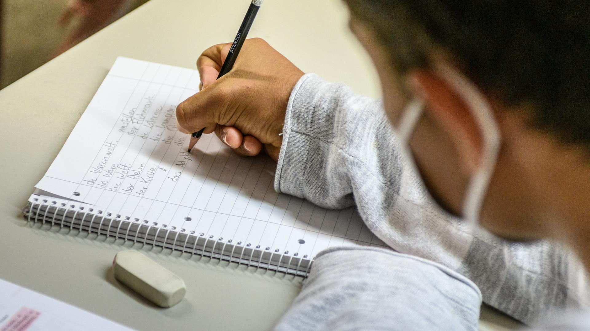 Ein junger Mann sitzt an einem Tisch und schreibt mit einem Bleistift auf einem linierten Spiralblock