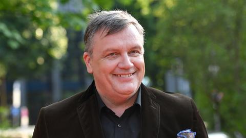 Porträtfoto eines lächelenden Mannes mit kurzen grauen Haaren. Er trägt ein dunkelbraunes Sakko über einem schwarzen Hemd. Im Hintergrund sind grüne Bäume zu sehen.