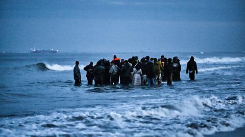 Geflüchtete stehen im Wasser des Ärmelkanals in Nordfrankreich. Sie sind relativ dick angezogen, was auf kalt Temperaturen hindeutet. Jugendliche und ggf. auch Kinder sind Teil der Gruppe.