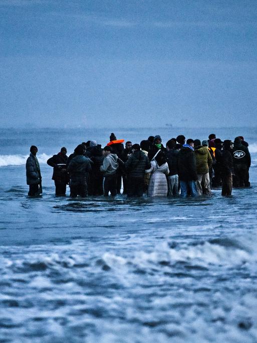 Geflüchtete stehen im Wasser des Ärmelkanals in Nordfrankreich. Sie sind relativ dick angezogen, was auf kalt Temperaturen hindeutet. Jugendliche und ggf. auch Kinder sind Teil der Gruppe. Geflüchtete stehen im Wasser des Ärmelkanals in Nordfrankreich. Sie sind relativ dick angezogen, was auf kalt Temperaturen hindeutet. Jugendliche und ggf. auch Kinder sind Teil der Gruppe.