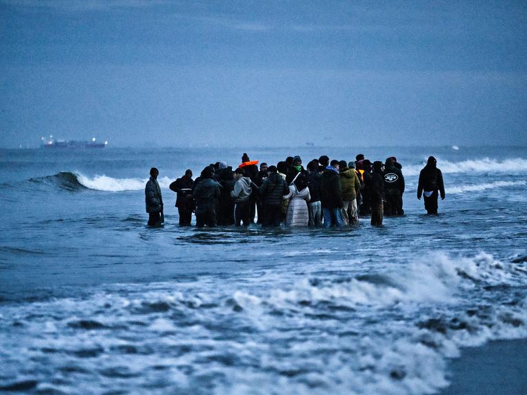 Geflüchtete stehen im Wasser des Ärmelkanals in Nordfrankreich. Sie sind relativ dick angezogen, was auf kalt Temperaturen hindeutet. Jugendliche und ggf. auch Kinder sind Teil der Gruppe. Geflüchtete stehen im Wasser des Ärmelkanals in Nordfrankreich. Sie sind relativ dick angezogen, was auf kalt Temperaturen hindeutet. Jugendliche und ggf. auch Kinder sind Teil der Gruppe.