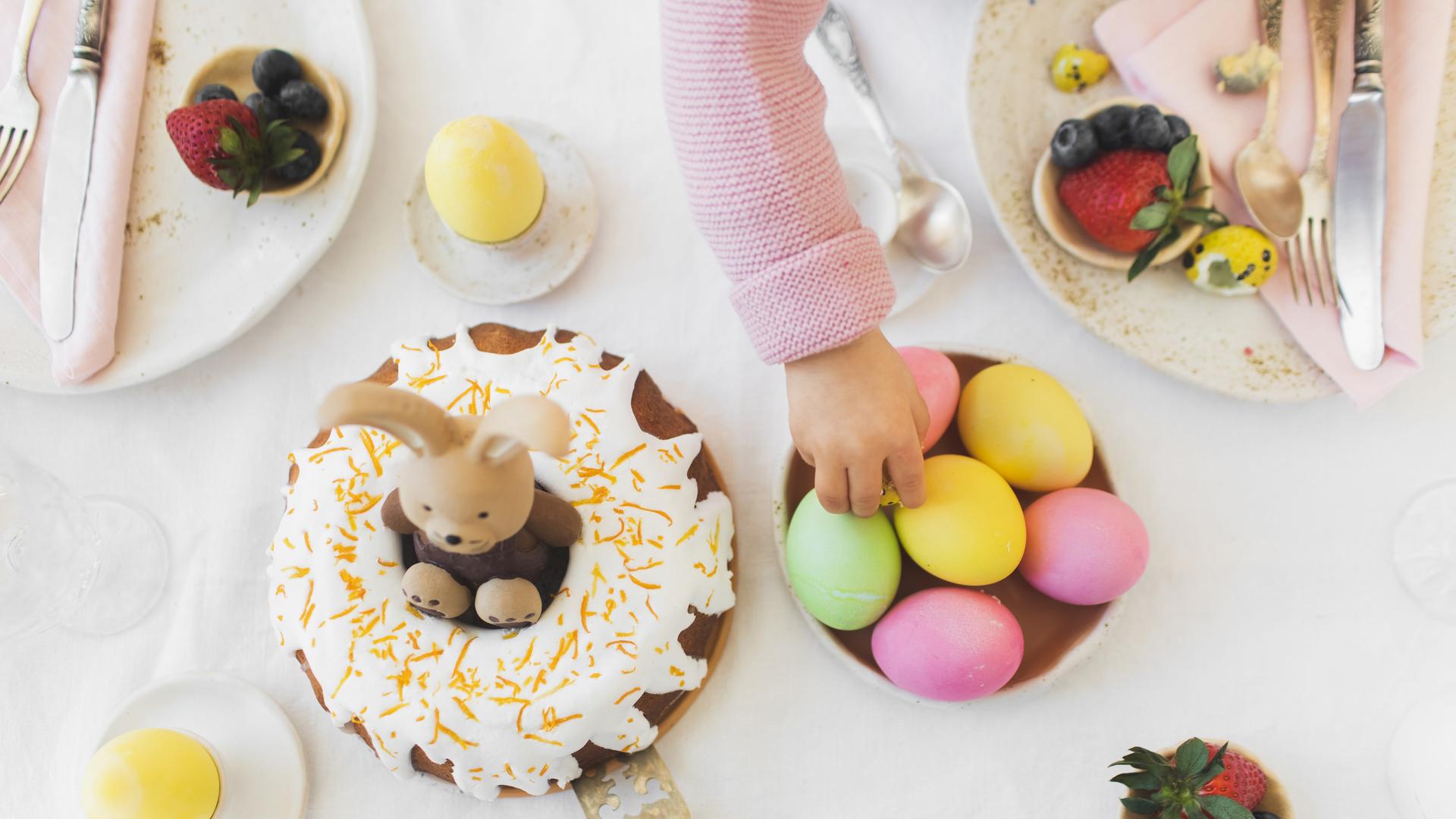 Ein für Ostern gedeckter und dekorierter Frühstückstisch. Eine kleine Kinderhand greift nach einem Ei.