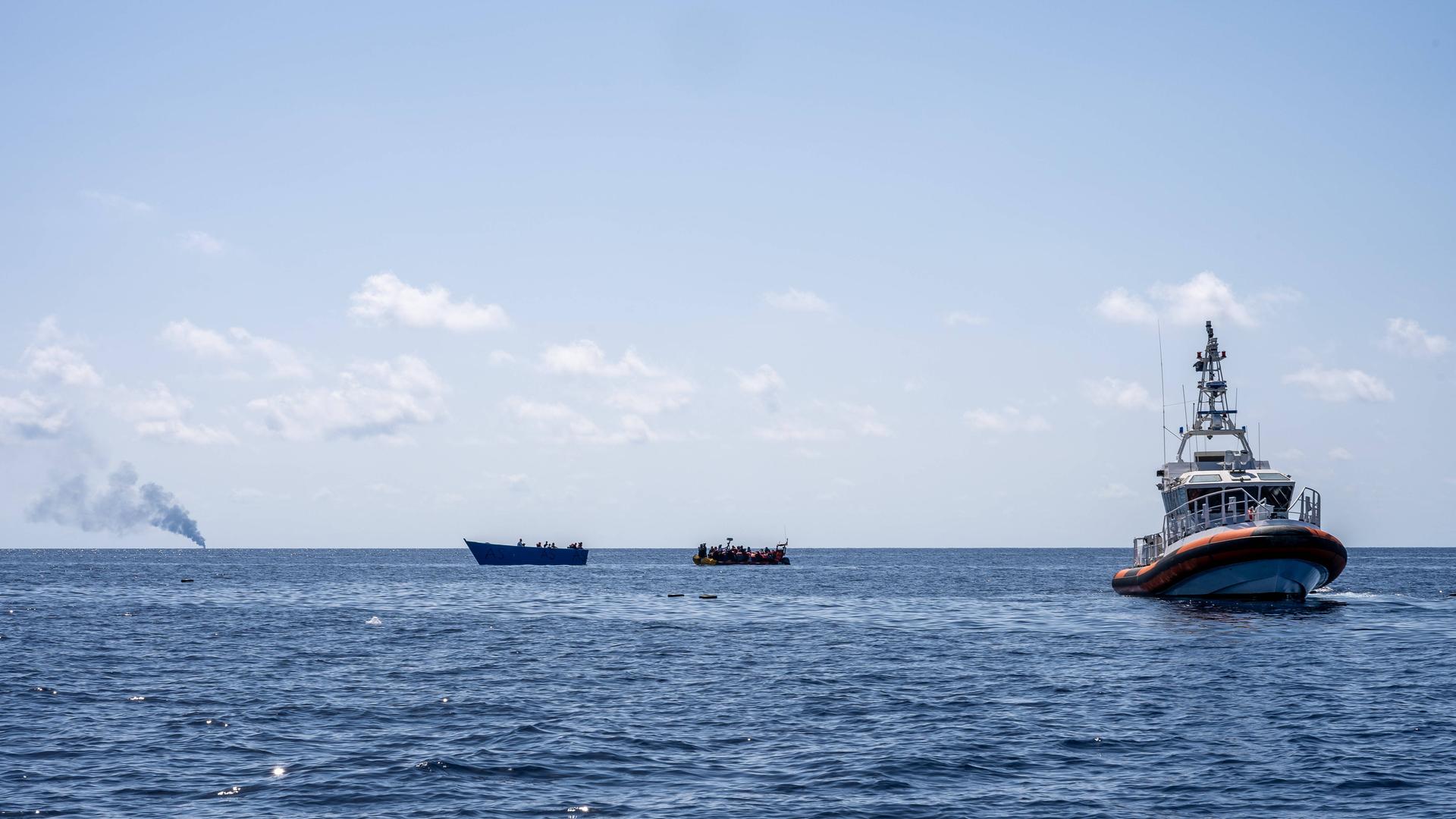 Auf dem offenen Meer sind links zwei kleine Boote mit Menschen zu erkennen, rechts ein größeres Boot der libyschen Küstenwache. Weiter hinten im Bild steigt Rauch auf.