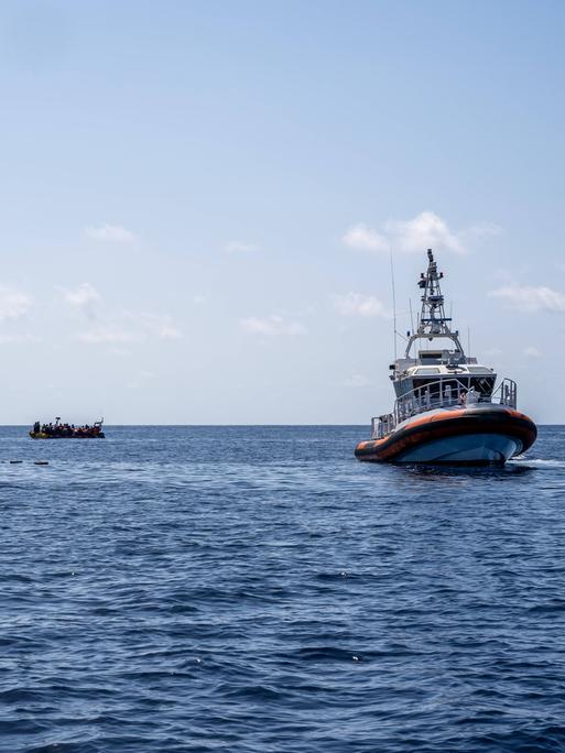 Auf dem offenen Meer sind links zwei kleine Boote mit Menschen zu erkennen, rechts ein größeres Boot der libyschen Küstenwache. Weiter hinten im Bild steigt Rauch auf.