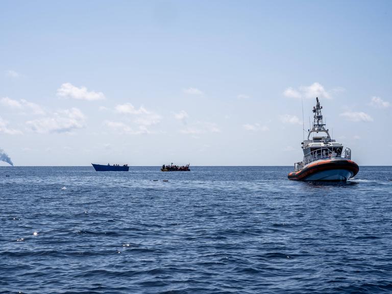 Auf dem offenen Meer sind links zwei kleine Boote mit Menschen zu erkennen, rechts ein größeres Boot der libyschen Küstenwache. Weiter hinten im Bild steigt Rauch auf.