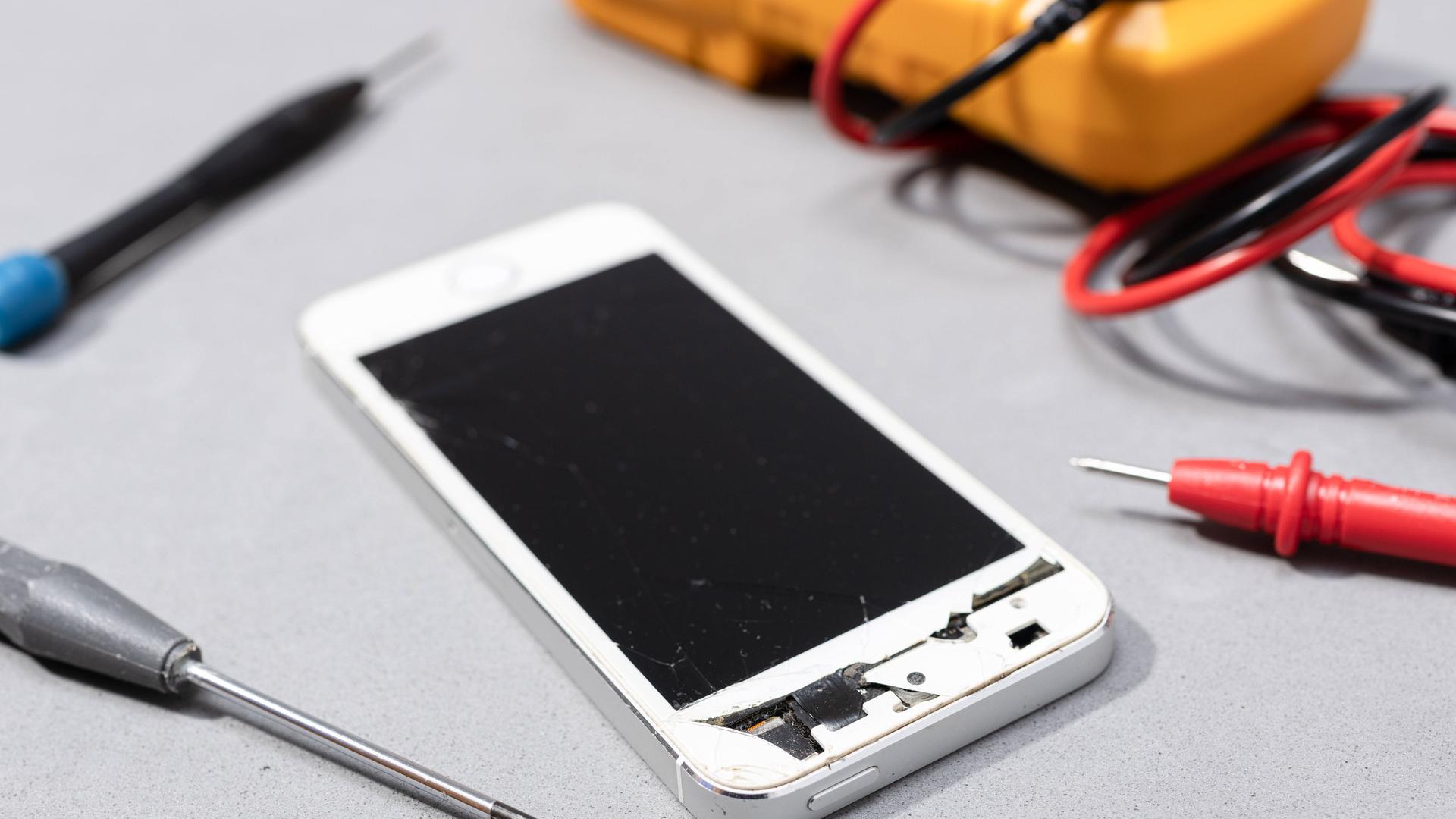 Reparatur von beschädigten Handy: Zu sehen ist ein Smartphone mit kaputtem Bildschirm auf der Werkbank eines Elektronikgeschäfts.