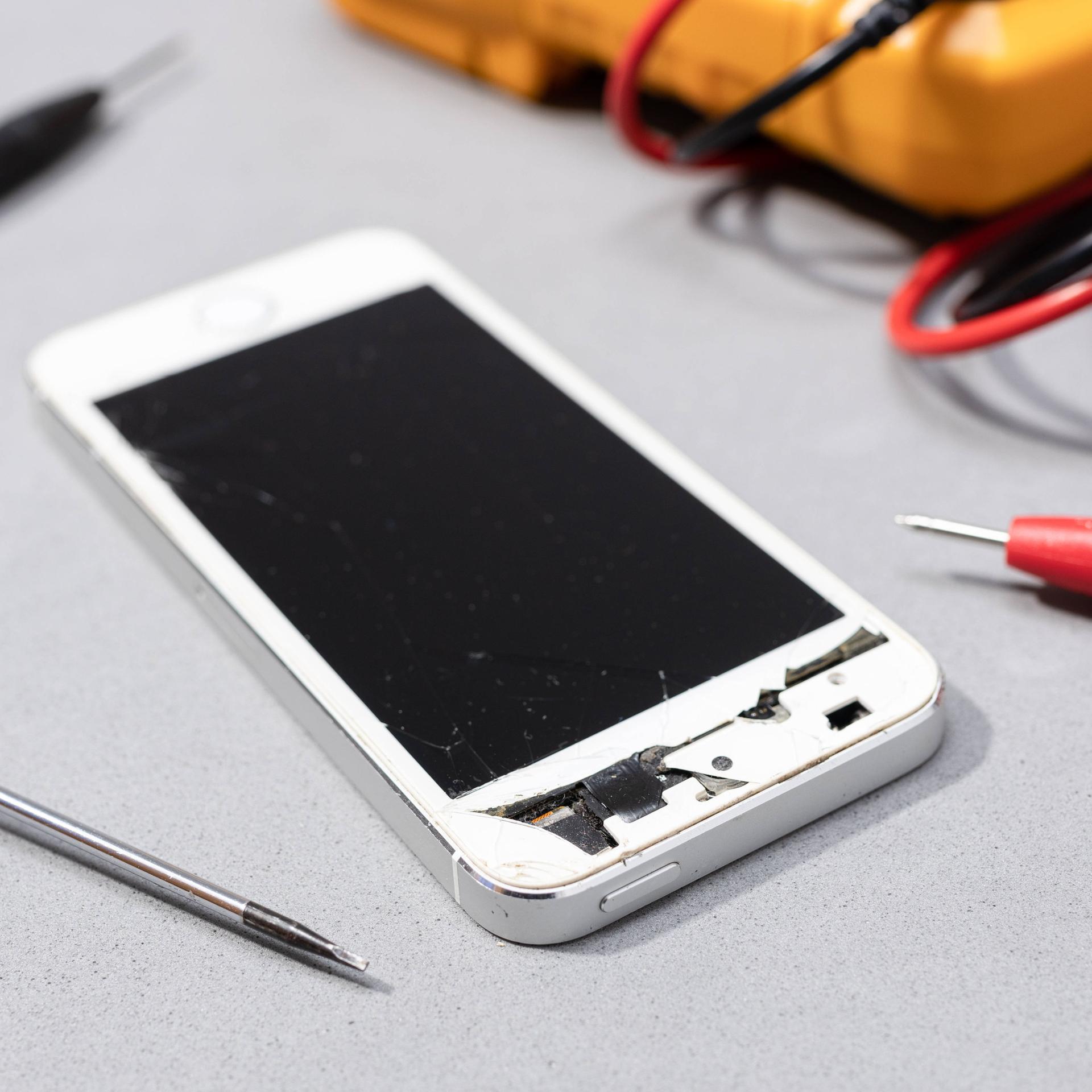Reparatur von beschädigten Handy: Zu sehen ist ein Smartphone mit kaputtem Bildschirm auf der Werkbank eines Elektronikgeschäfts.