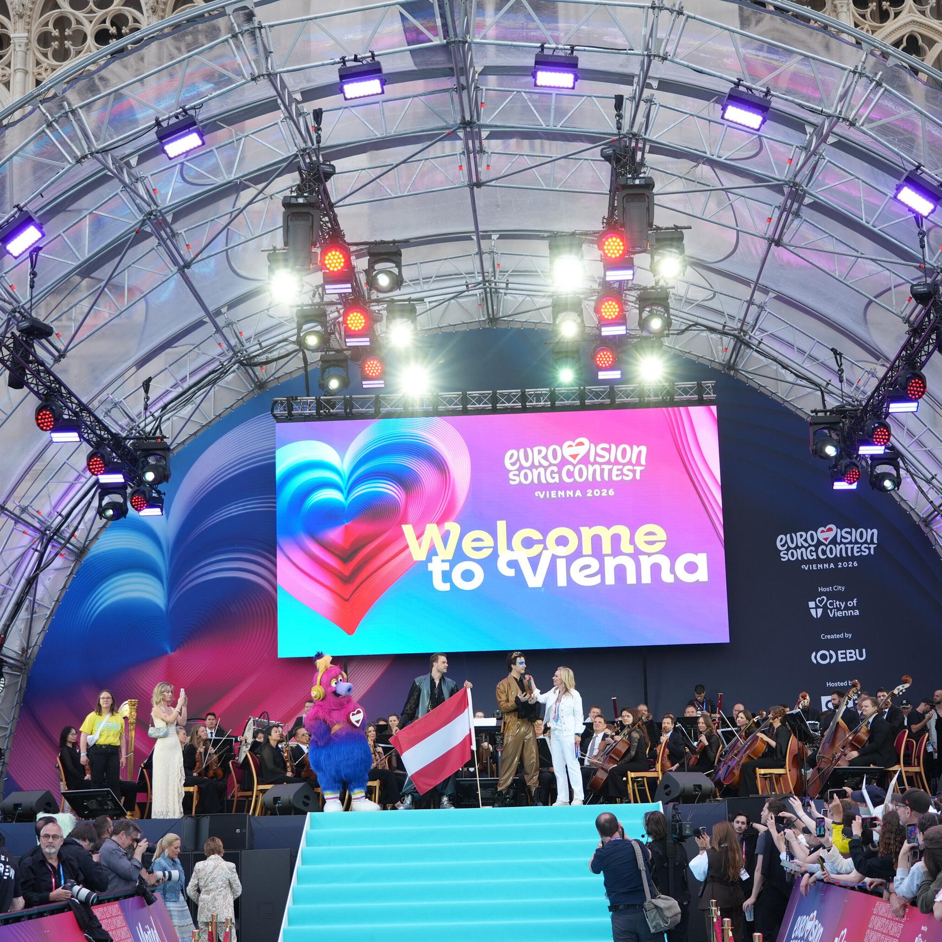 Die Eröffnungsfeier der ESC-Woche in Wien - Moderatoren und Sänger stehen auf der Bühne. Unten sind entlang eines Laufstegs Publikum und Kamerateams zu sehen.
