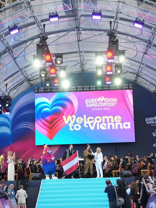 Die Eröffnungsfeier der ESC-Woche in Wien - Moderatoren und Sänger stehen auf der Bühne. Unten sind entlang eines Laufstegs Publikum und Kamerateams zu sehen.