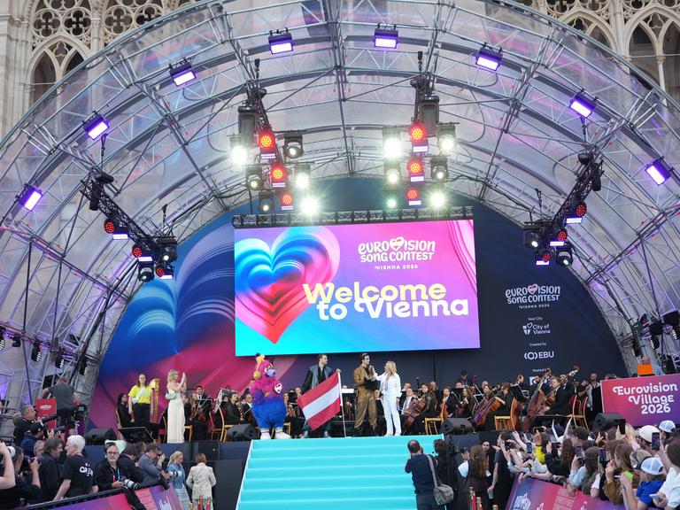 Die Eröffnungsfeier der ESC-Woche in Wien - Moderatoren und Sänger stehen auf der Bühne. Unten sind entlang eines Laufstegs Publikum und Kamerateams zu sehen.