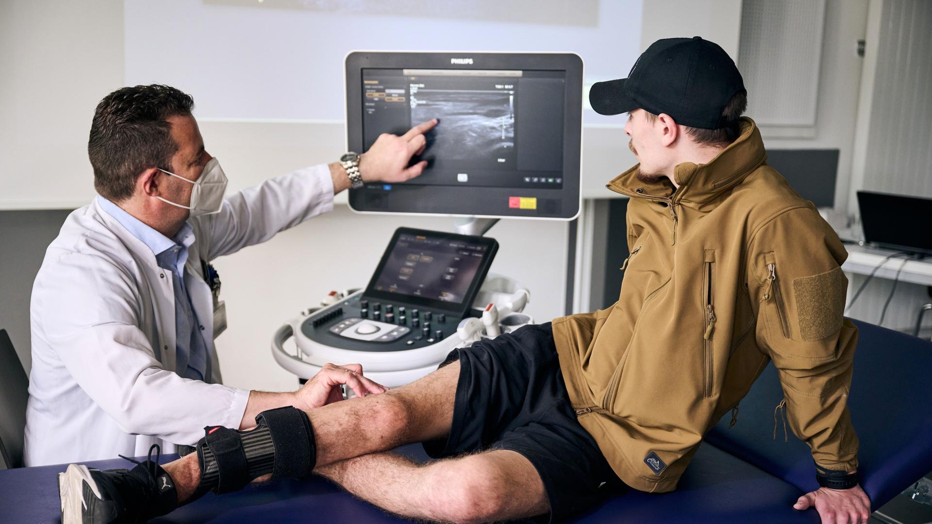 Ein am Bein verletzter Mann sitzt auf einer Liege und wird von einem Arzt untersucht. Ein am Bein verletzter Mann sitzt auf einer Liege und wird von einem Arzt untersucht.