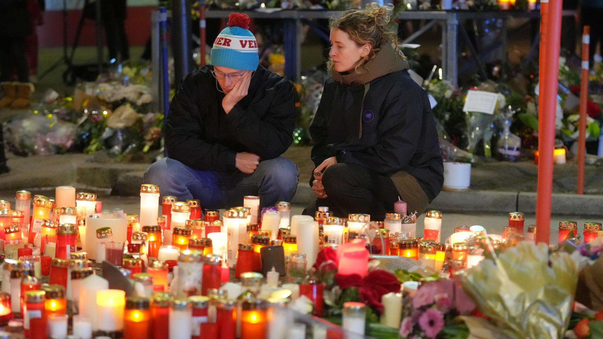 Ein Mann und eine Frau sitzen traurig vor vielen Kerzen und Blumen. 