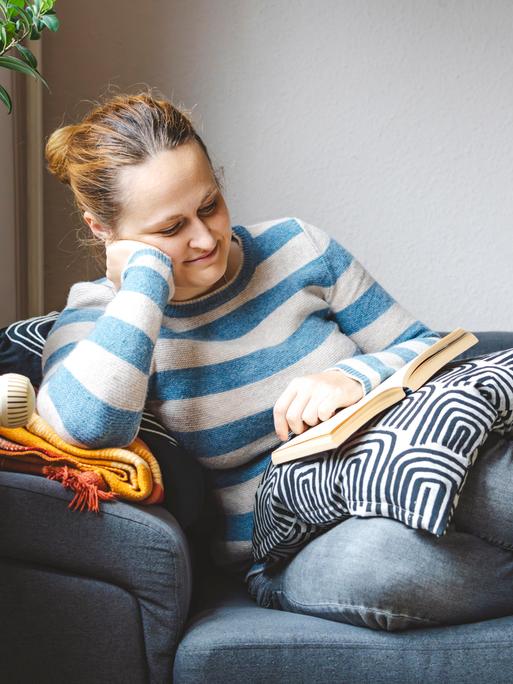Buch lesende Frau auf Sofa. Sie hat einen Pullover an, es sieht gemütlich aus. 